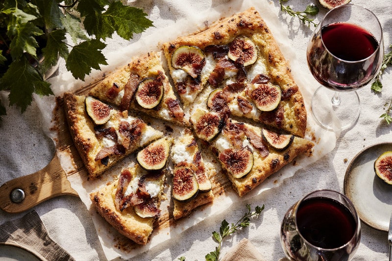 Flammkuchen mit Feige und Ricotta auf mediterranem Tisch, dazu ein Glas Rotwein – perfekt kombiniert mit Wein aus AustralienFlammkuchen mit Feige und Ricotta auf mediterranem Tisch, dazu ein Glas Rotwein – perfekt kombiniert mit Wein aus Australien