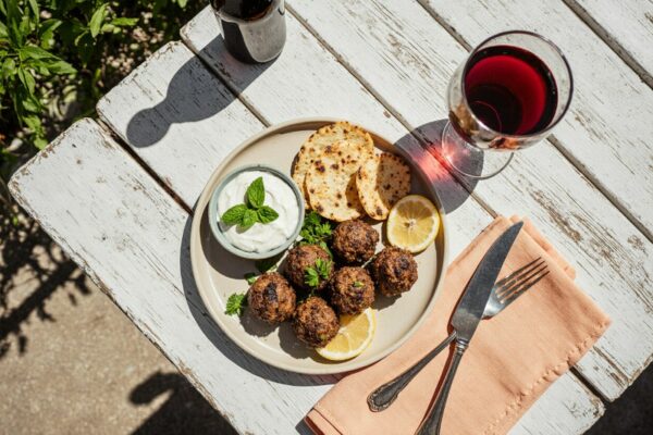 Lammbällchen mit Minzjoghurt auf weißem Teller, Rotweinglas und Flasche daneben – serviert auf sommerlicher Terrasse mit Grillambiente und einem Glas Wein aus Chile