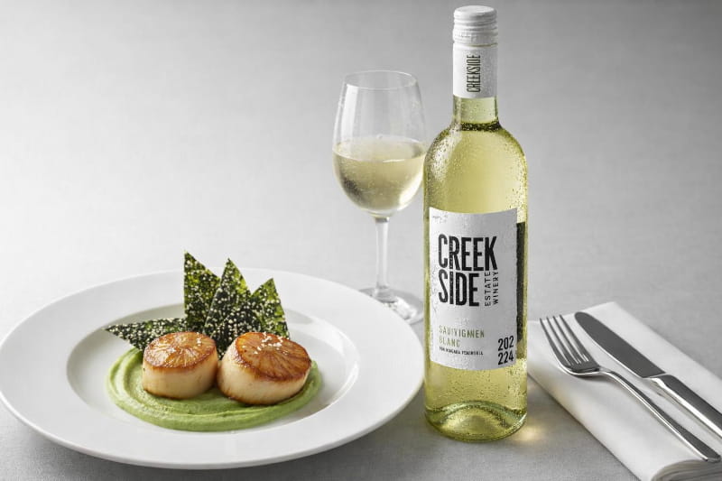 Zwei goldbraune Jakobsmuscheln auf smaragdgrüner Erbsen-Wasabi-Creme, daneben knusprige Nori-Chips, auf weißem Teller mit Fleur de Sel, rechts ein Weißweinglas mit hellgelbem Sauvignon Blanc, leicht beschlagen
