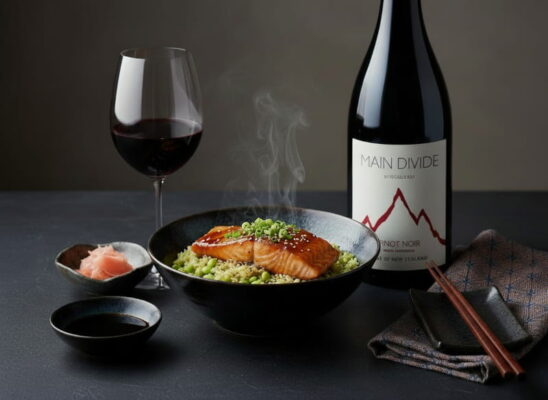 Teriyaki-Lachs mit lackglänzender Glasur auf Edamame-Reis in einer japanischen Schüssel, daneben eingelegter Ingwer und ein Rotweinglas mit leuchtendem Pinot Noir, schwarzer Sesam als Dekoration