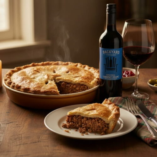 Goldbraune Tourtière auf rustikalem Holztisch, angeschnitten mit sichtbarer Hackfleischfüllung, daneben ein Rotweinglas mit tiefem Rubinrot, Preiselbeersoße und Cornichons als Beilage