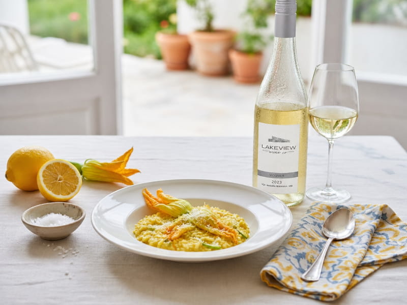 Cremiges Zucchiniblüten-Risotto mit leuchtend gelben Blüten, Parmesanspänen und Zitronenabrieb in einem tiefen weißen Teller, daneben ein schlankes Weißweinglas mit hellgoldenem Viognier, frische Zitrone und Zucchiniblüten als Dekoration