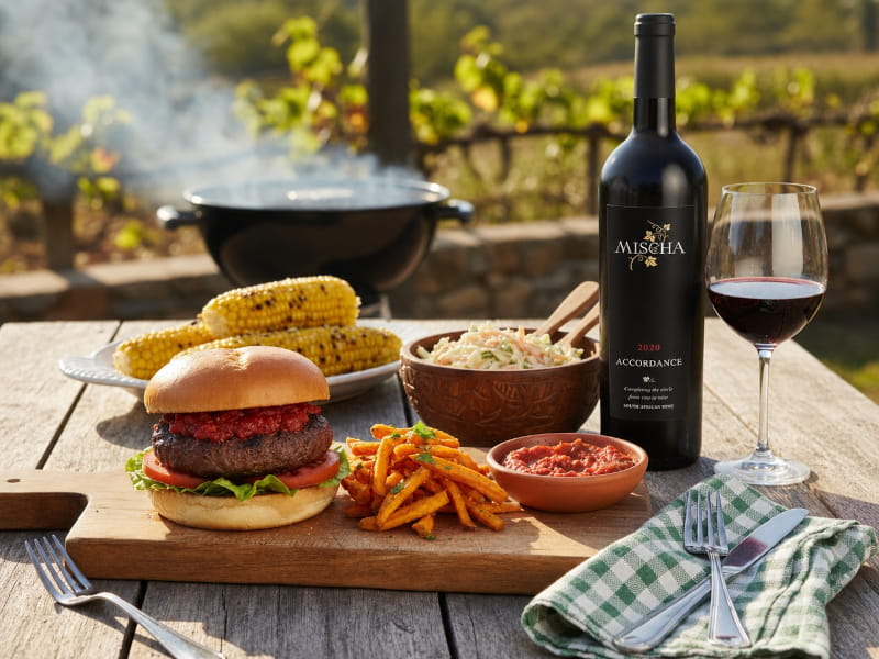 Südafrikanischer Braai-Burger mit Chakalaka-Relish und Süßkartoffelchips auf rustikalem Holzbrett, daneben ein Glas Accordance von Mischa Estate aus Wellington, Südafrika