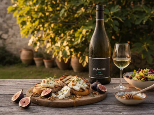 Gegrilltes Brot mit Gorgonzola, Feigen und Walnüssen auf einem Holzbrett, daneben Honig, Salat und ein Glas Elephant Hill Sea Viognier aus Hawke's Bay, Neuseeland, im mediterranen Garten mit Feigenbaum im Hintergrund